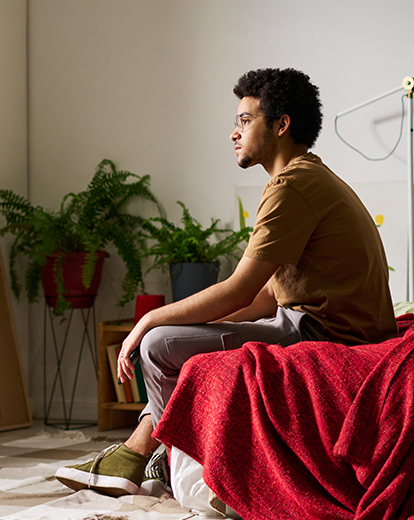 Junger Mann sitzt auf seinem Bett und schaut nachdenklich aus dem Fenster