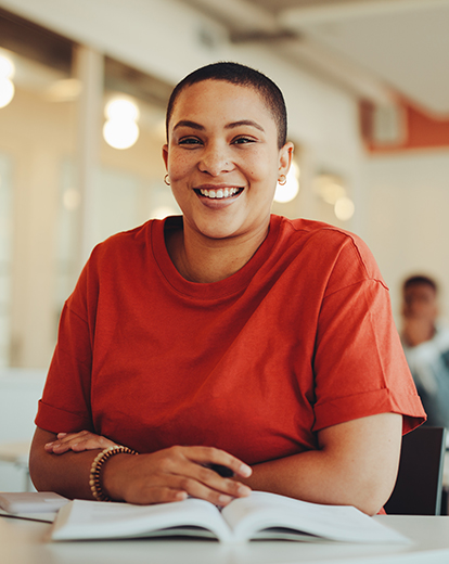 Junge Frau vor einem Buch in einem Klassenraum grinst 