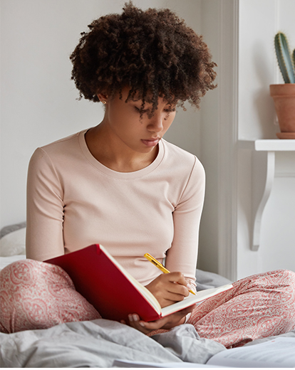 Eine junge Frau sitzt auf einem Bett und schreibt in ein Buch.
