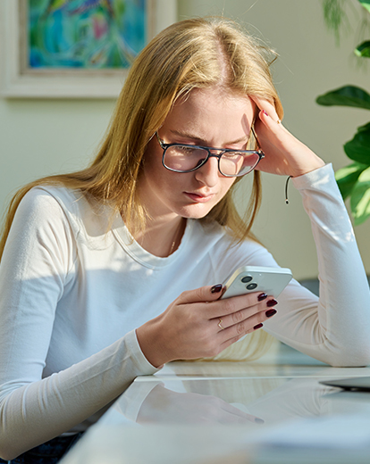 Junge Frau stützt Kopf in die Hände und schaut nachdenklich in ihr Telefon
