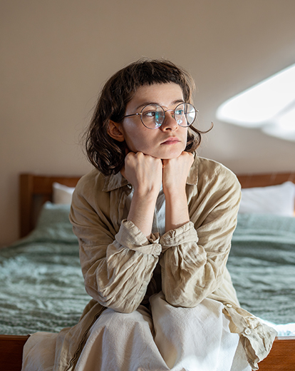 Junge Frau sitzt auf ihrem Bett und hat nachdenklich ihren Kopf auf ihre Fäuste gestützt