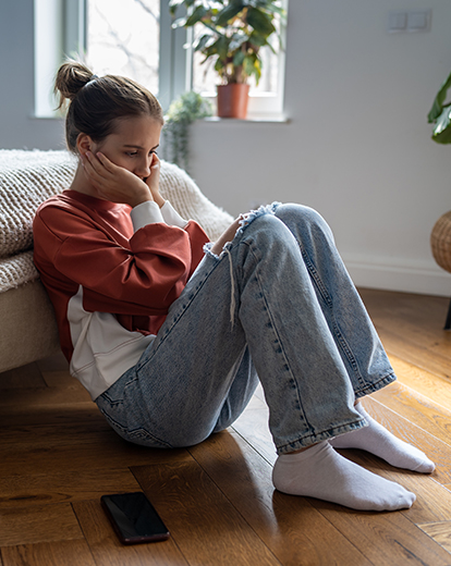 Junge Frau sitzt auf dem Boden vor ihrem Bett und hat verzweifelt ihren Kopf in ihre Hände gestützt