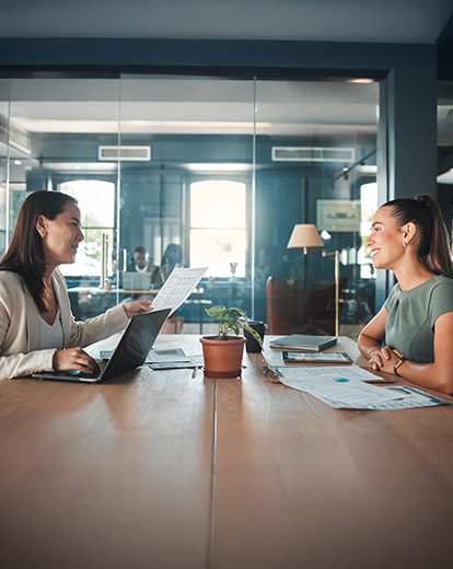 Junge Frau sitzt an einem Tisch und unterhält sich mit älterer Frau, die Unterlagen in der Hand hat