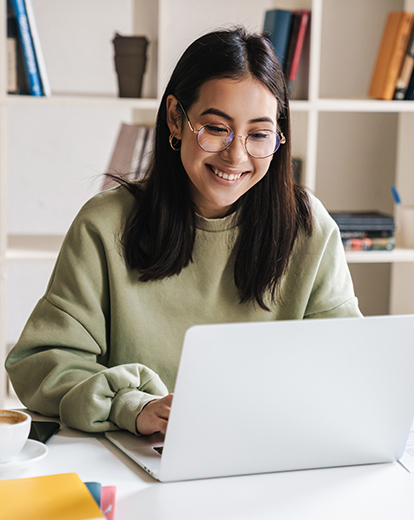 Junge Frau sitzt lächelnd vor ihrem Laptop an ihrem Arbeitsplatz