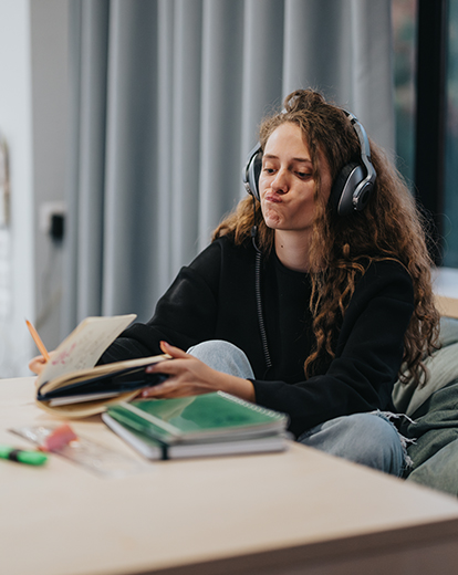 Junge Studentin sitzt auf einem Sofa mit Kopfhörern und schreibt in ein Buch