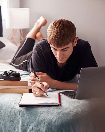 Student liegt auf einem Bett vor seinem Laptop und schreibt auf ein Papier