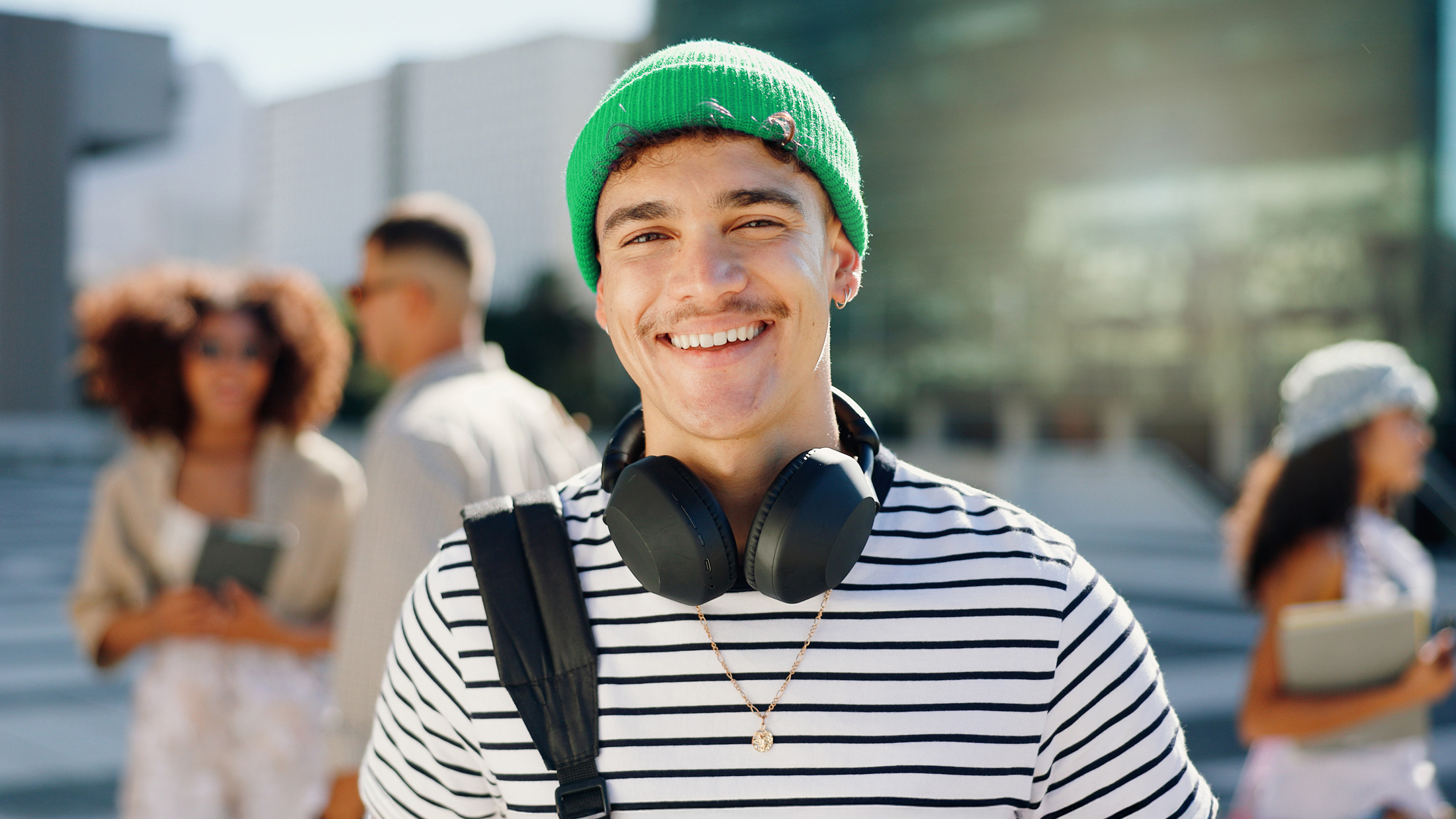 Student mit grüner Beanie und Over-Ear-Kopfhörern lächelt entspannt in die Kamera, im Hintergrund ein belebter Campus bei Tageslicht.